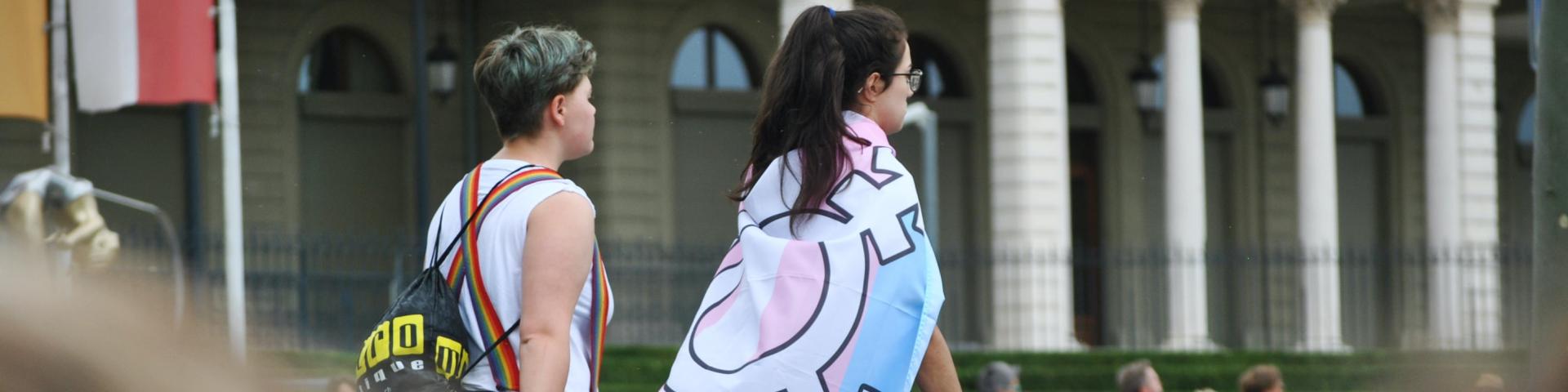 Two people walk along a road, one wrapped in a PRIDE flag with the trans inclusive colours, as part of a PRODE march 