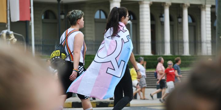 Two people walk along a road, one wrapped in a PRIDE flag with the trans inclusive colours, as part of a PRODE march 