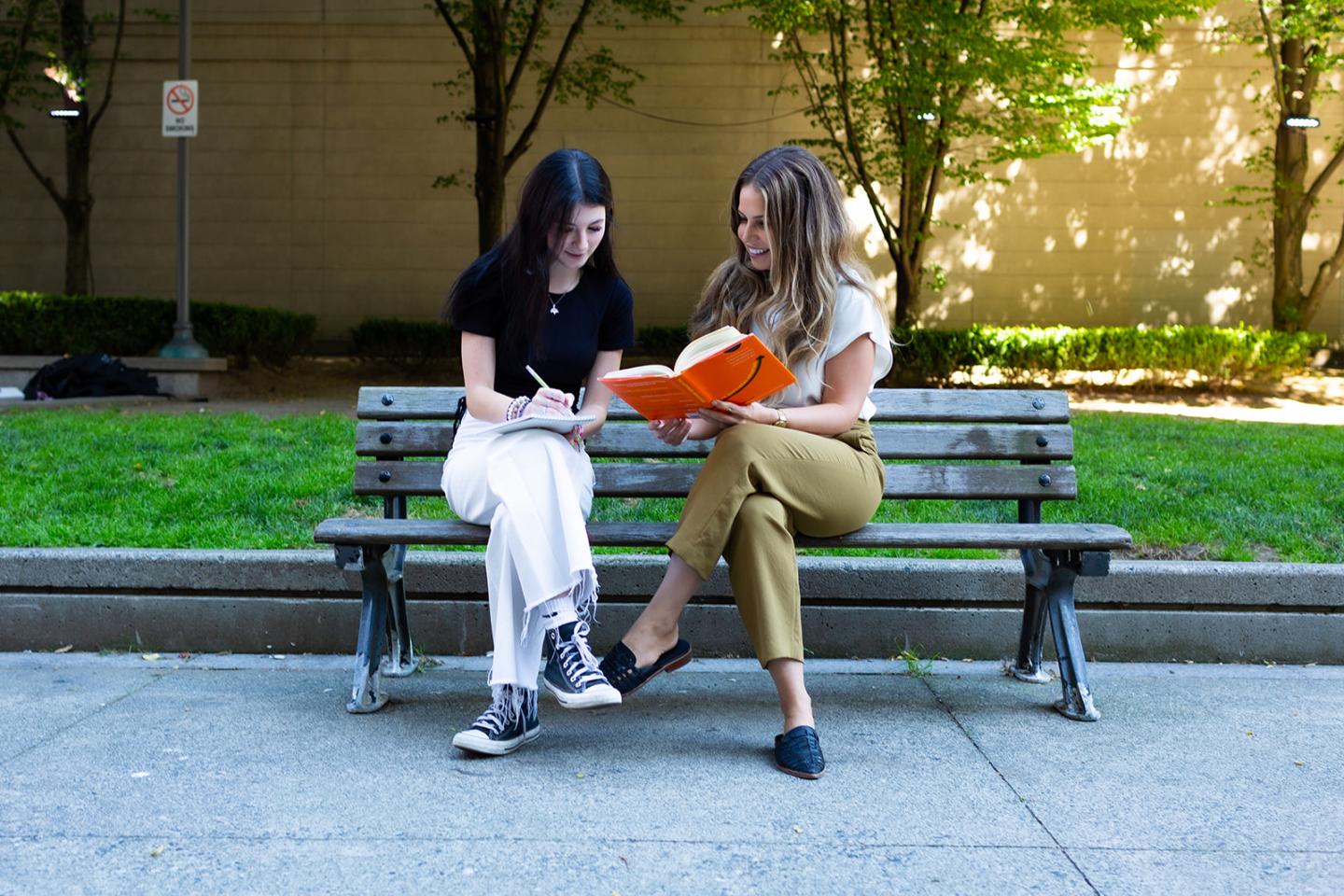 Young female sitting on a bench. On the left side the mentee wearing a black shirt, white pants is taking notes while her mentor reads a orange cover book