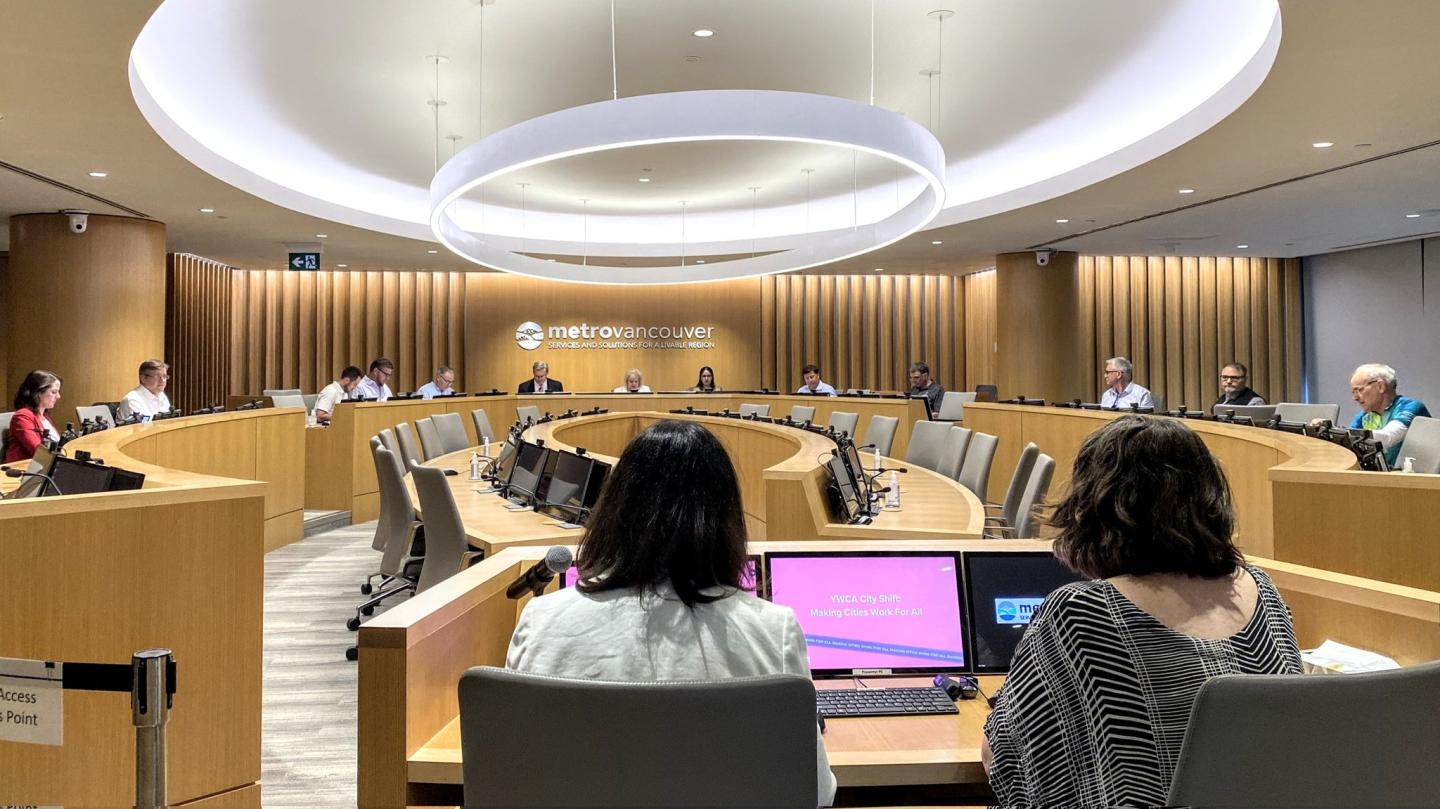 Women looking at two screens facing a room with people sitting around a circle. 