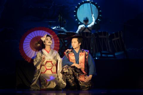 Two performers in traditional Japanese dress kneel on stage, with a Taiko drum being played in the background