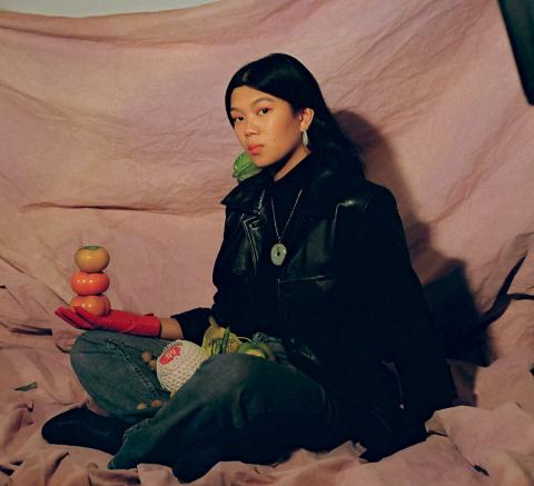 Portrait of a woman sitting with a pink-beige background, holding fruits in her hands.