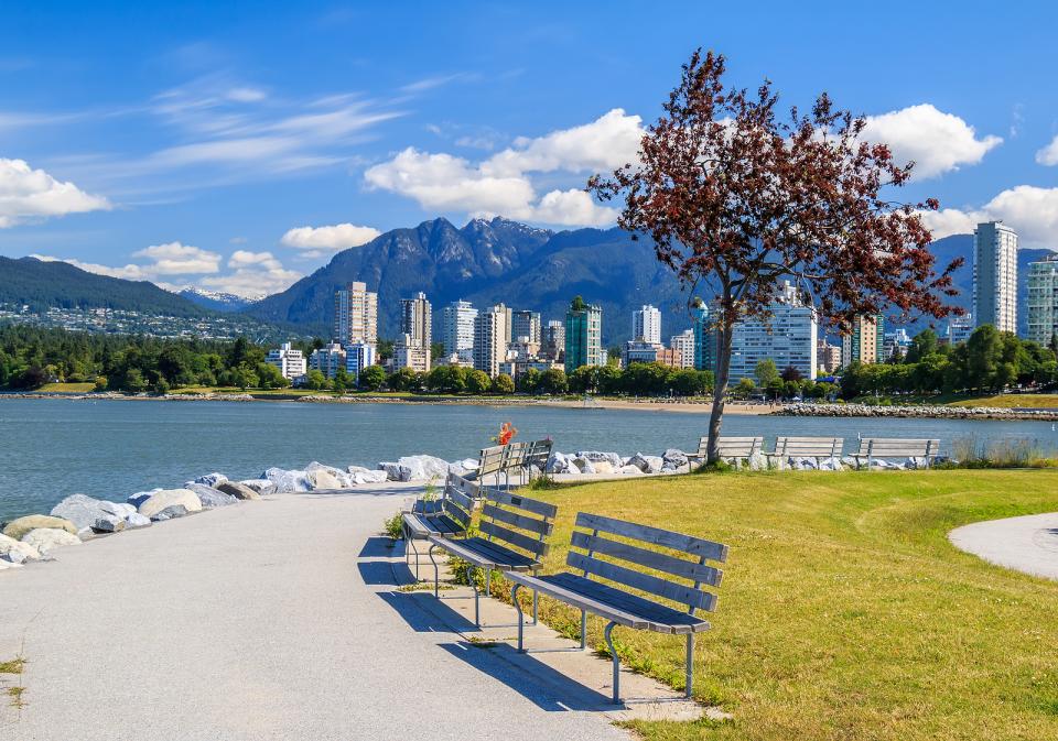 Benches, grass, tree on along the curved path of the West End sea wall overlooking the skyline of mountains and blue skies