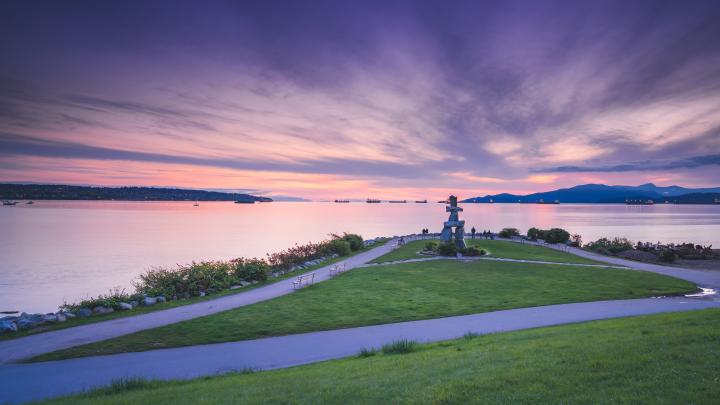 A field and the Inukshuk at English Bay in Vancouver with a dazzling sky of purple, orange, pink and different colours at dusk.